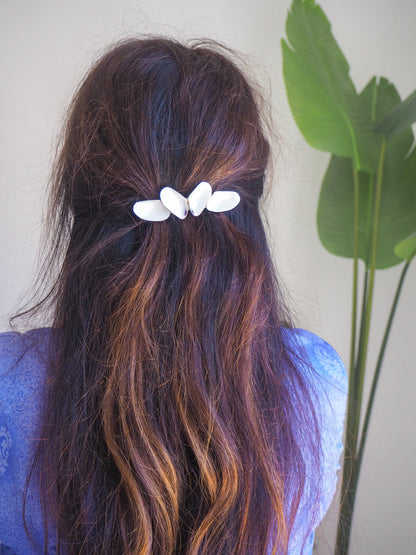 Person with wavy hair wearing a white hair clip, standing next to a plant. Model wearing White Coquina Shells Butterfly Hair Clip Barrette, Handpicked Real Coquina Shells. The shells are white in color and attached on the hair clip like a butterfly. The model has brown long hair. Sea Butterfly Shell Hair Decoration, Seashell Wedding Hair accessories, Coastal Christmas gift, Beach Wedding Hair