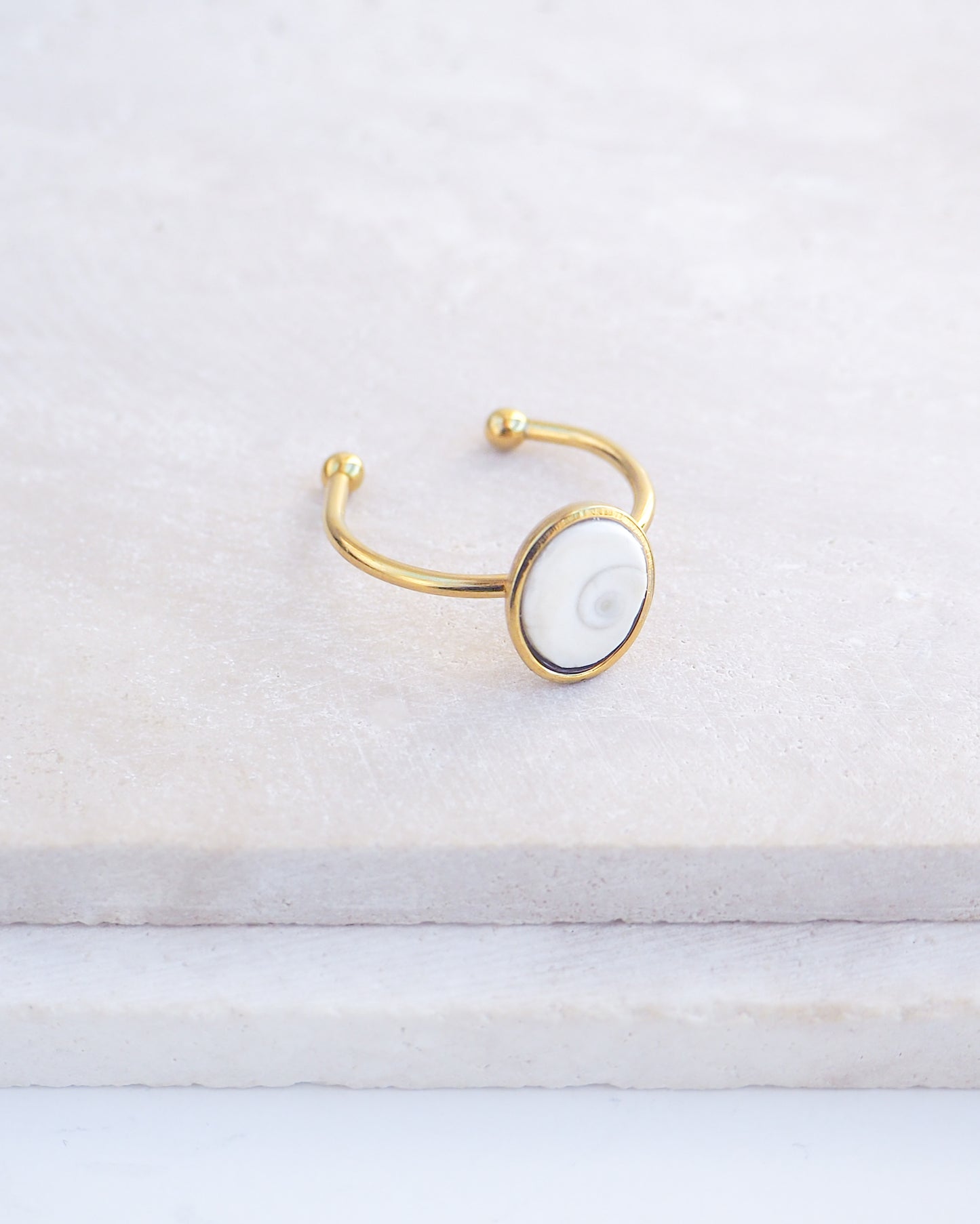 Side view of Gold bezel ring with a tiny ivory white shiva eye on stone displayed with light background. White Eye of Saint Lucia Gold Ring on stone displayed, 18k gold stainless steel shiva eye operculum, Saint Lucy Eye, operculum pendant