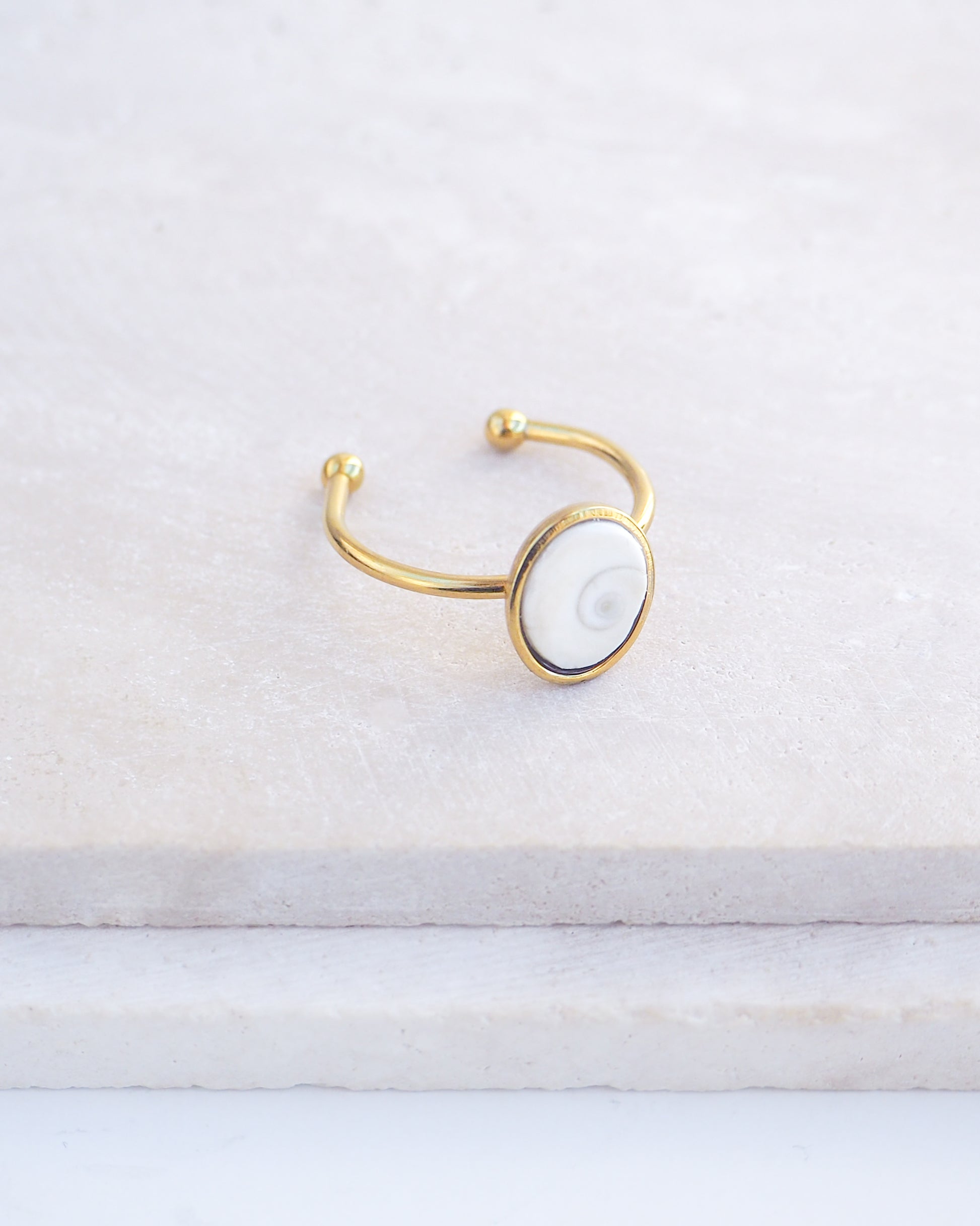 Side view of Gold bezel ring with a tiny ivory white shiva eye on stone displayed with light background. White Eye of Saint Lucia Gold Ring on stone displayed, 18k gold stainless steel shiva eye operculum, Saint Lucy Eye, operculum pendant