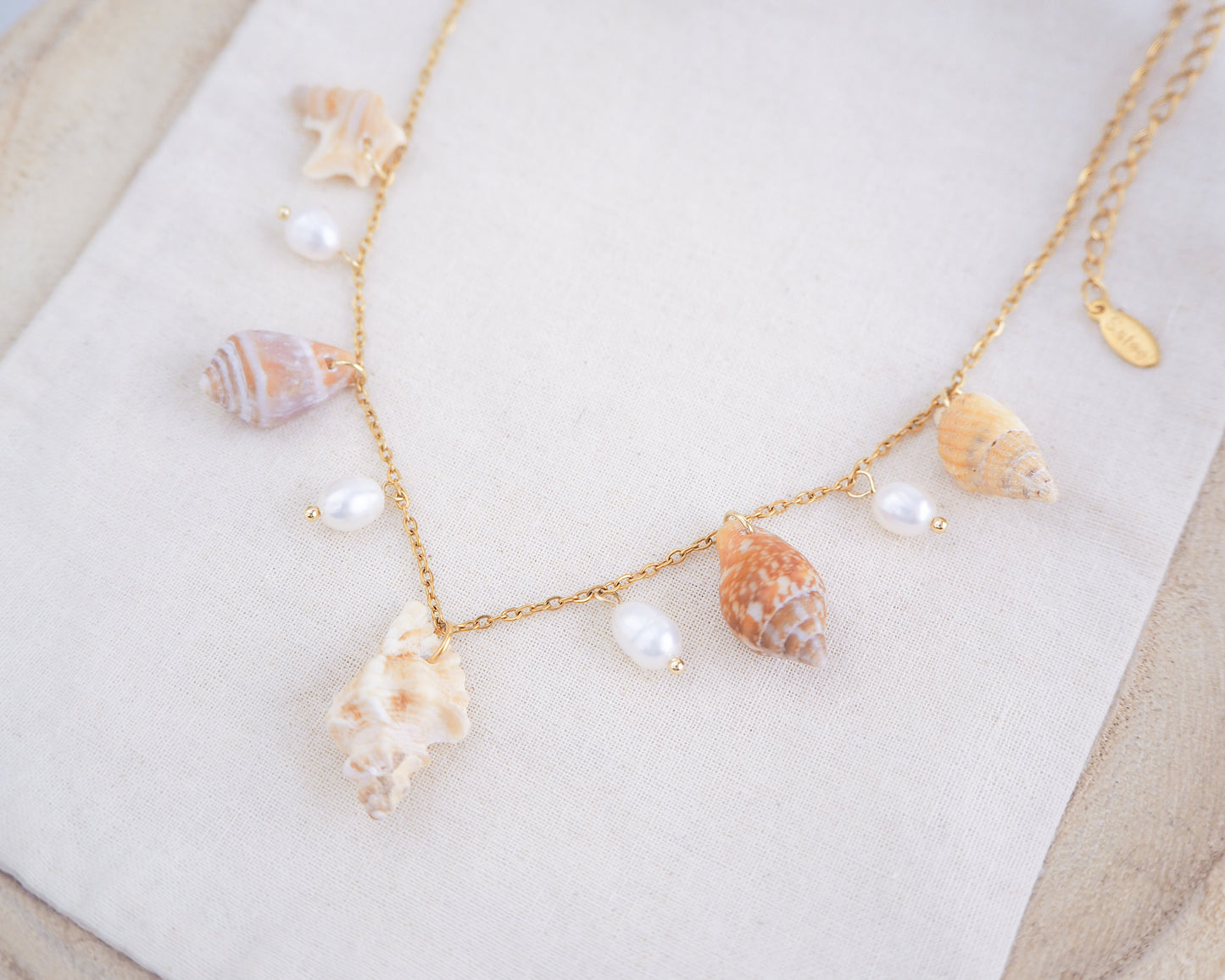 Side view of necklace with seashells and pearls on a white surface. Portuguese Shell Pearl Charm Necklace with tiny shells from Portugal. Mediterranean cone shell, pelican foot shell, rustic dove shell, netted dog whelk shell, sting winkle shell. Ivory white Freshwater pearls