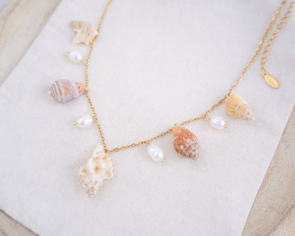 Side view of necklace with seashells and pearls on a white surface. Portuguese Shell Pearl Charm Necklace with tiny shells from Portugal. Mediterranean cone shell, pelican foot shell, rustic dove shell, netted dog whelk shell, sting winkle shell. Ivory white Freshwater pearls