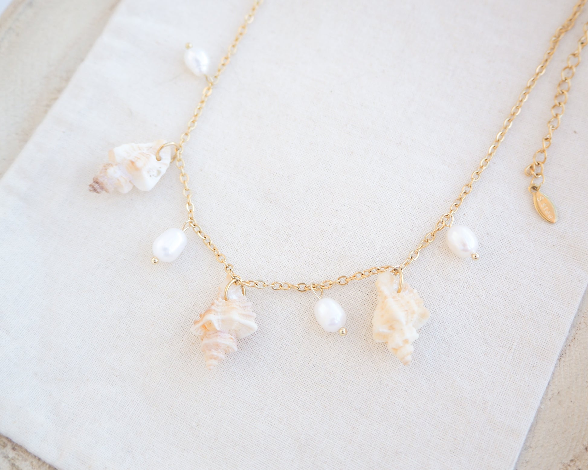 Side view of gold necklace with seashells and pearls on cotton. Portuguese Shell Pearl Charm Necklace with tiny sting winkle shells from Portugal and ivory white Freshwater pearls.