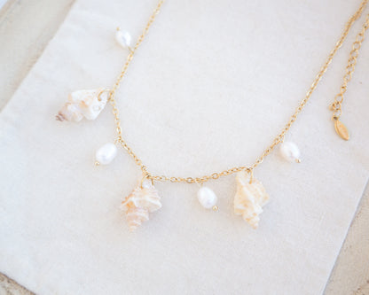 Side view of gold necklace with seashells and pearls on cotton. Portuguese Shell Pearl Charm Necklace with tiny sting winkle shells from Portugal and ivory white Freshwater pearls.
