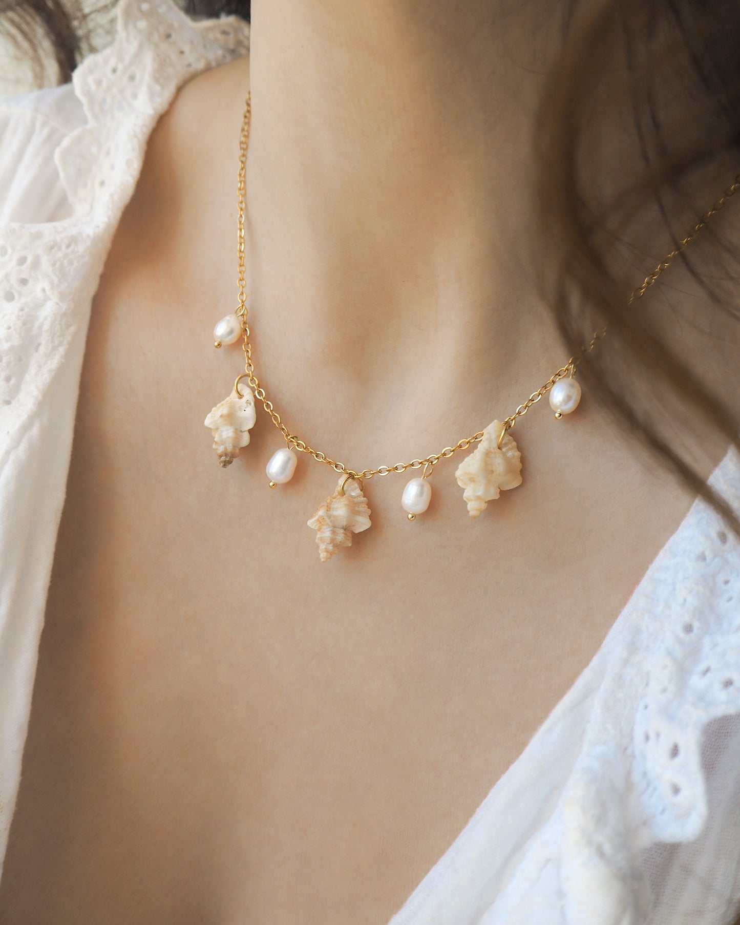 Model wearing a gold necklace with seashells and pearls on neck. Portuguese Shell Pearl Charm Necklace with tiny sting winkle shells from Portugal and ivory white Freshwater pearls.