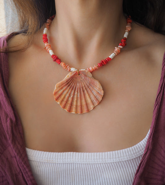 Model wearing a necklace with a big scallop shell paired with red stone beads, pink stone beads and white shell beads. The shell is a great scallop shell from Portugal and is about 7 cm. The color of the shell is orange pink and it has vertical ridges. 