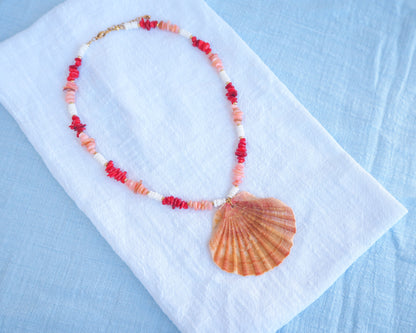 Necklace with red and white beads and a shell pendant on a white cloth with a blue background. Necklace with shell pendant and red and white beads on a light blue background. Big scallop shell necklace paired with red stone beads, pink stone beads and white shell beads. The shell is a great scallop shell from Portugal and is about 7 cm. The color of the shell is orange pink and it has vertical ridges. 