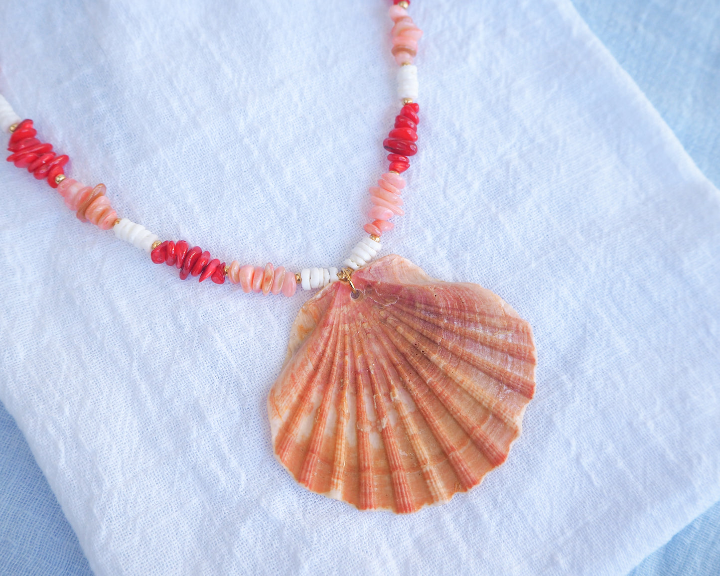 Necklace with shell pendant and red and white beads on a light blue background. Big scallop shell necklace paired with red stone beads, pink stone beads and white shell beads. The shell is a great scallop shell from Portugal and is about 7 cm. The color of the shell is orange pink and it has vertical ridges. 
