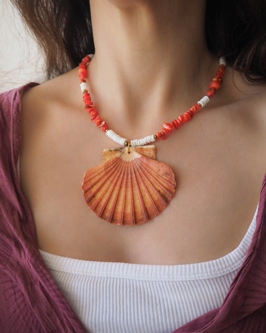 Necklace with a large shell pendant and coral beads worn by a person. Model wearing a necklace with a big scallop shell paired with red stone beads and white shell beads. The shell is a great scallop shell from Portugal and is about 8 cm. The color of the shell is orange pink and it has vertical ridges. 