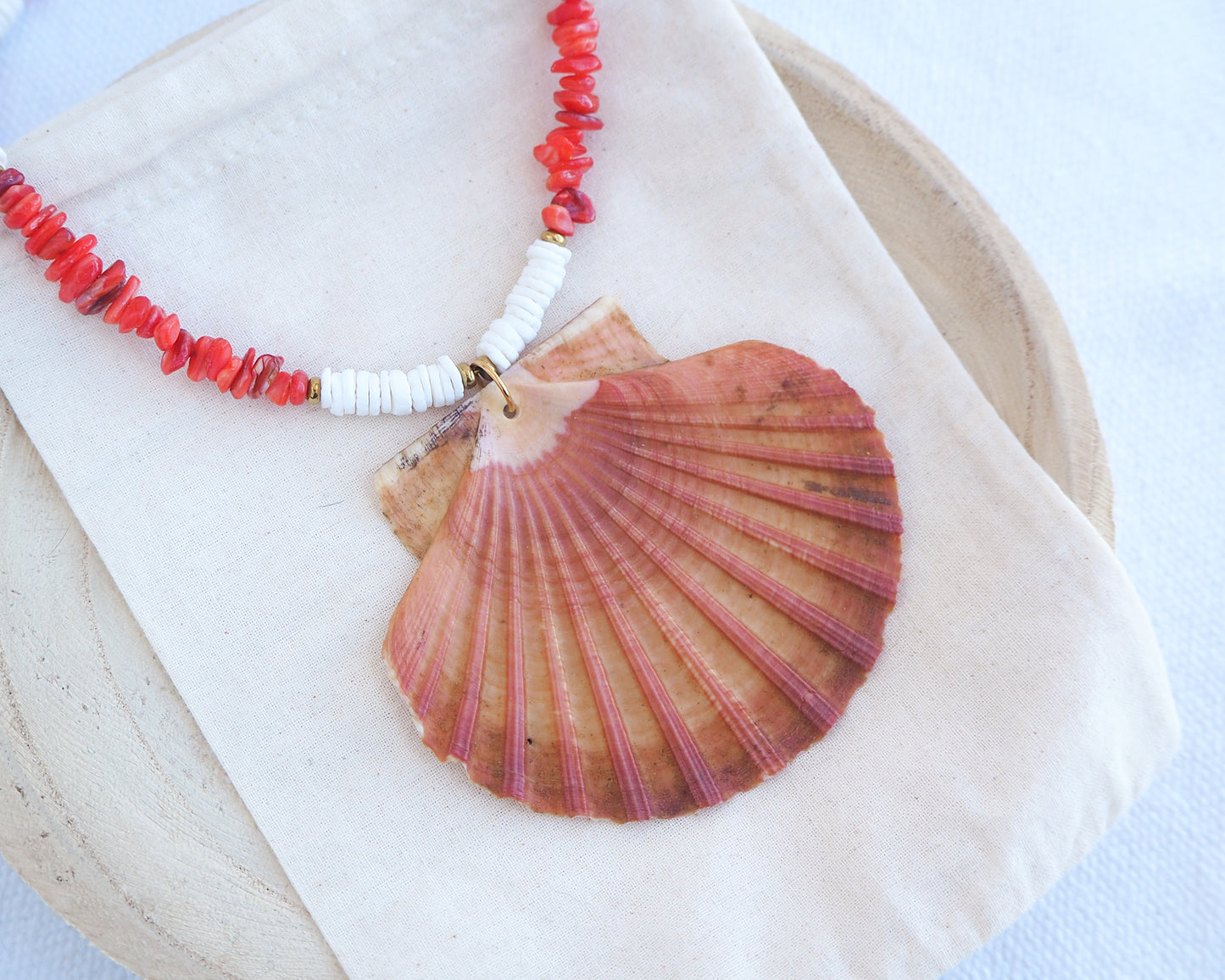 Side view of a necklace with a large shell pendant and red beads worn by a person. Model wearing a necklace with a big scallop shell paired with red stone beads and white shell beads. The shell is a great scallop shell from Portugal and is about 8 cm. The color of the shell is orange pink and it has vertical ridges. 