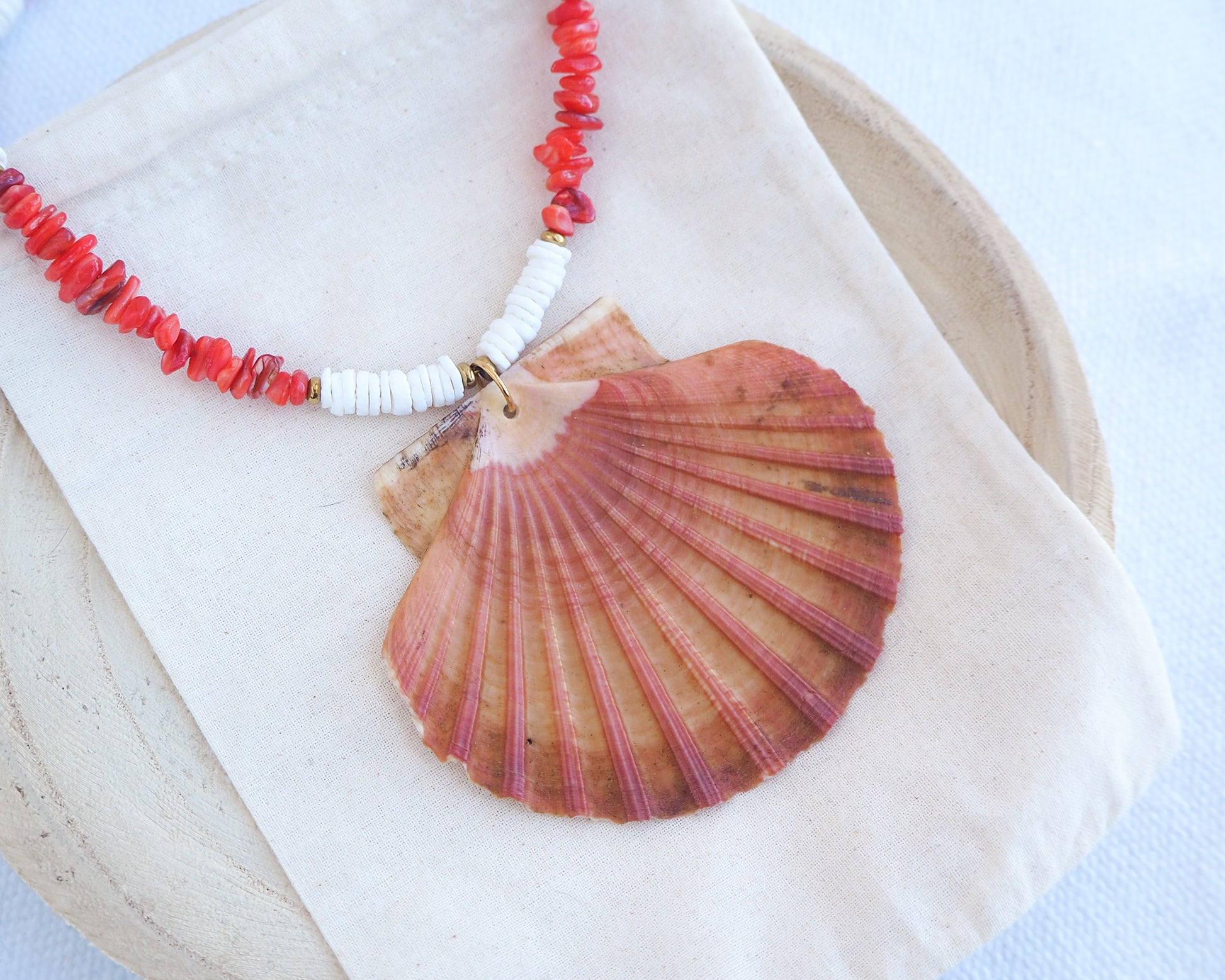 Side view of a necklace with a large shell pendant and red beads worn by a person. Model wearing a necklace with a big scallop shell paired with red stone beads and white shell beads. The shell is a great scallop shell from Portugal and is about 8 cm. The color of the shell is orange pink and it has vertical ridges. 