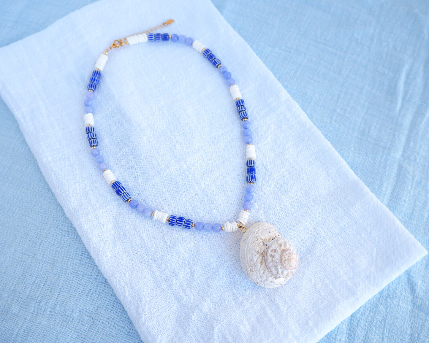 Necklace with blue and white beads and a shell pendant on a light blue fabric background. A handmade necklace with a big star shell paired with blue angelite gemstone beads, blue striped beads and white shell beads. The shell is a rough star shell from Portugal and is about 5 cm. The color of the shell is ivory white and round in shape.