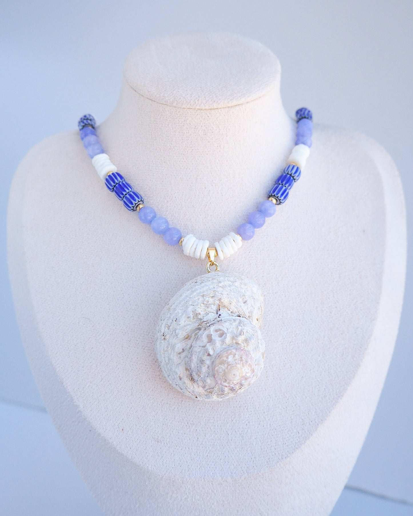 Necklace with a large round pendant on a mannequin bust against a light blue background. A handmade necklace with a big star shell paired with blue angelite gemstone beads, blue striped beads and white shell beads. The shell is a rough star shell from Portugal and is about 5 cm. The color of the shell is ivory white and round in shape.