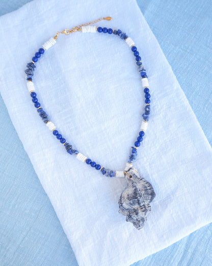 a handmade necklace with a big grey murex shell paired with blue sodalite gemstone beads and white shell beads. The shell is a banded dye murex shell from Portugal and is about 8 cm. The color of the shell is grey.
