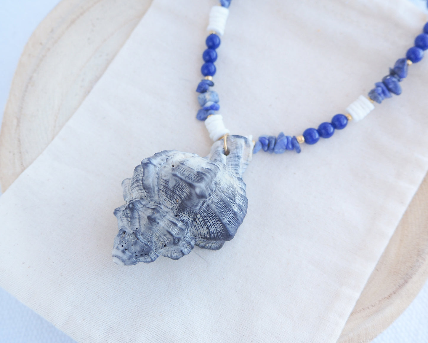 Side view of a handmade necklace with a big grey murex shell paired with blue sodalite gemstone beads and white shell beads. The shell is a banded dye murex shell from Portugal and is about 8 cm. The color of the shell is grey.
