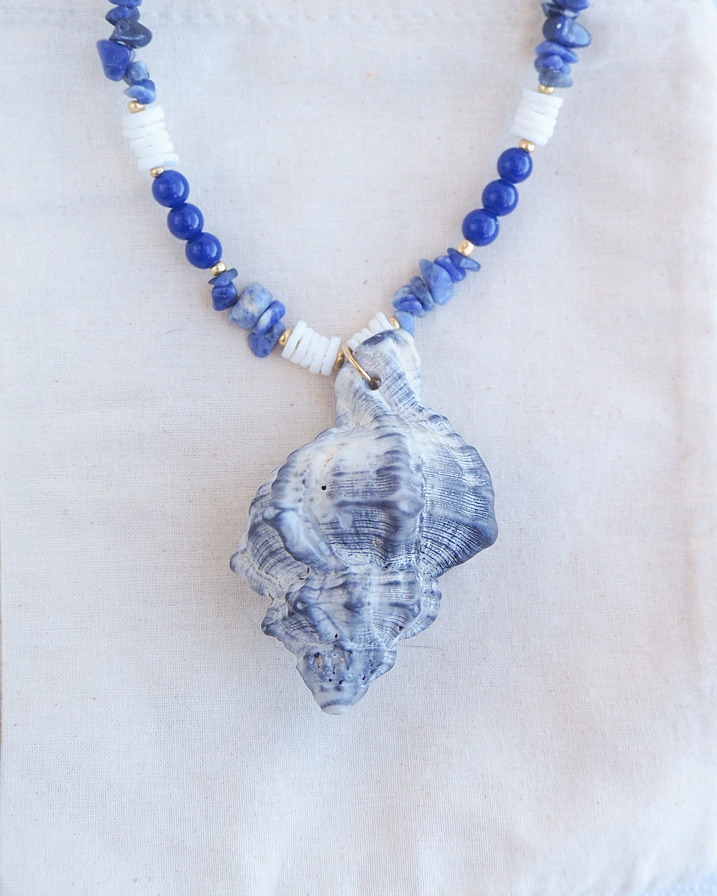 Front view close up of handmade necklace with a big grey murex shell paired with blue sodalite gemstone beads and white shell beads. The shell is a banded dye murex shell from Portugal and is about 8 cm. The color of the shell is grey.