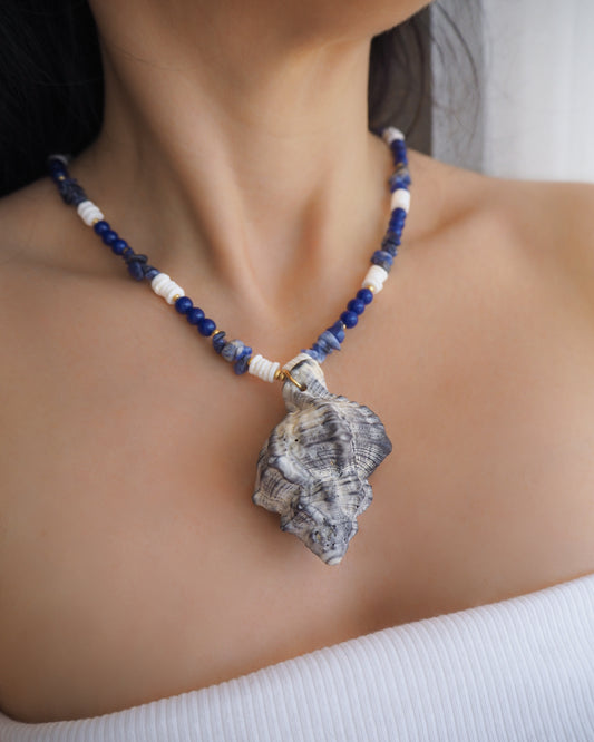Model wearing a necklace with a big grey murex shell paired with blue sodalite gemstone beads and white shell beads. The shell is a banded dye murex shell from Portugal and is about 8 cm. The color of the shell is grey.
