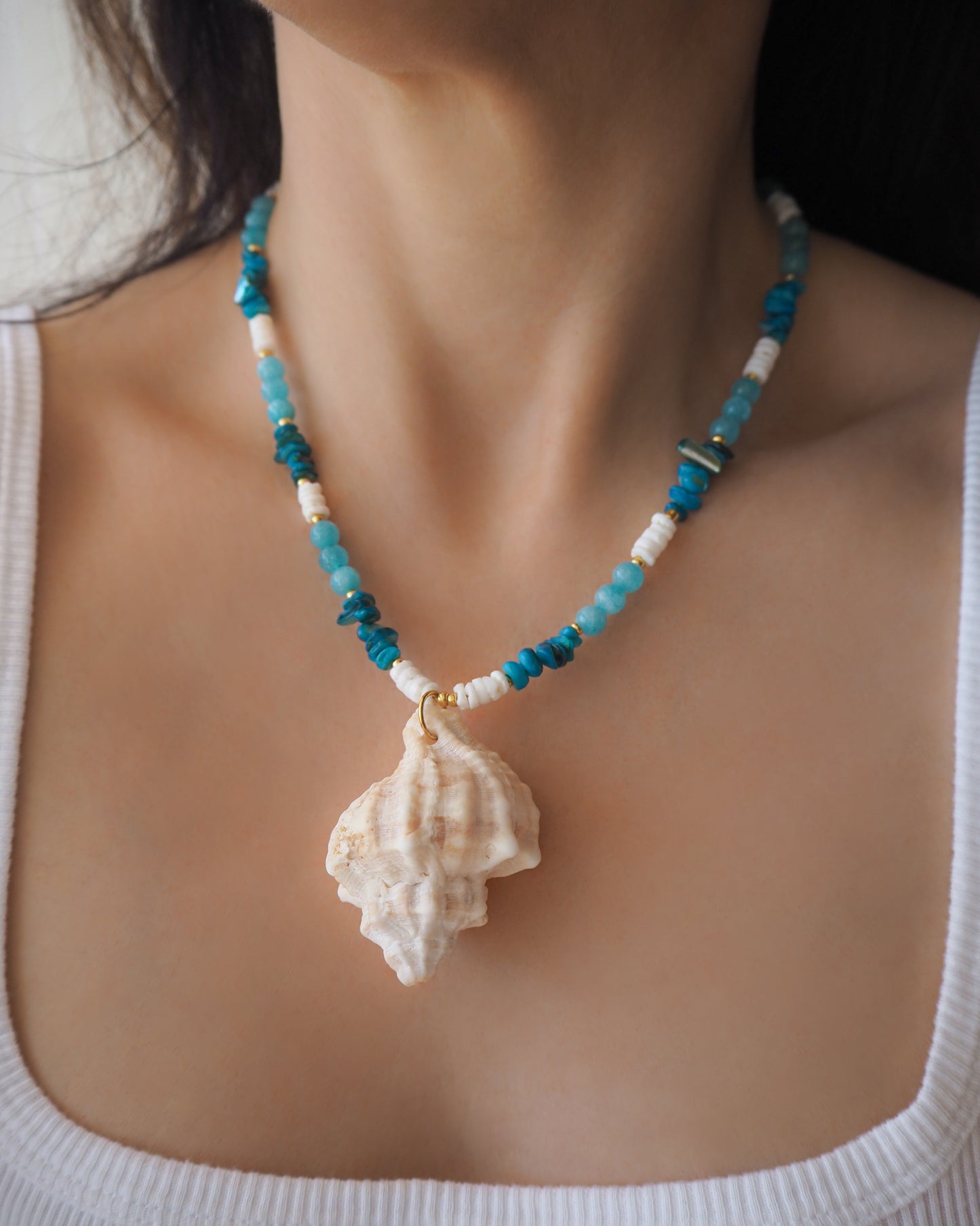 Model wearing a necklace with a big murex shell paired with aquamarine gemstone beads and white shell beads. The shell is a banded dye murex shell from Portugal and is about 6 cm. The color of the shell is ivory bege.