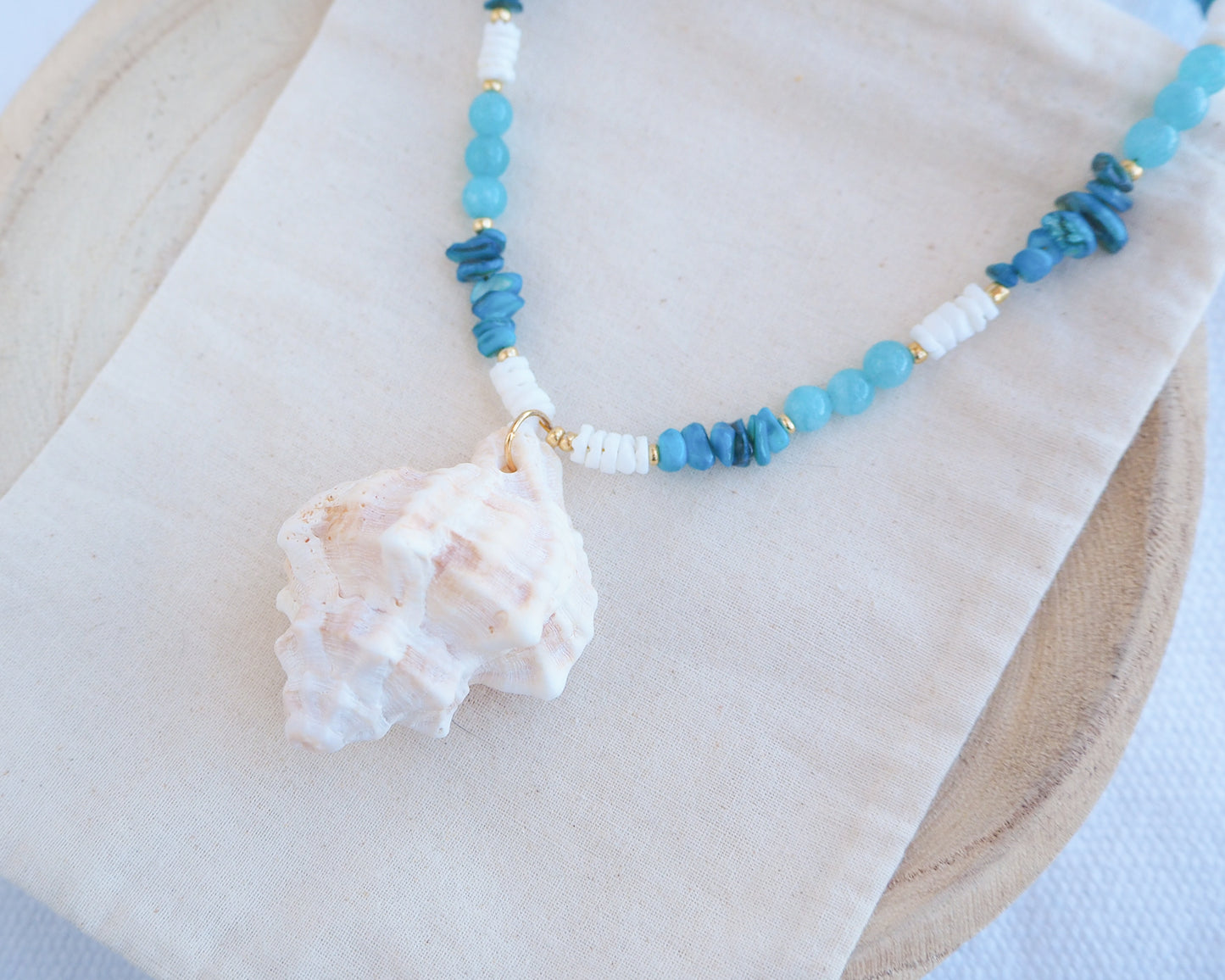 Necklace with blue beads and a shell pendant on a white surface. A necklace with a ivory murex shell paired with aquamarine gemstone beads and white shell beads. The shell is a banded dye murex shell from Portugal and is about 6 cm. The color of the shell is ivory bege.