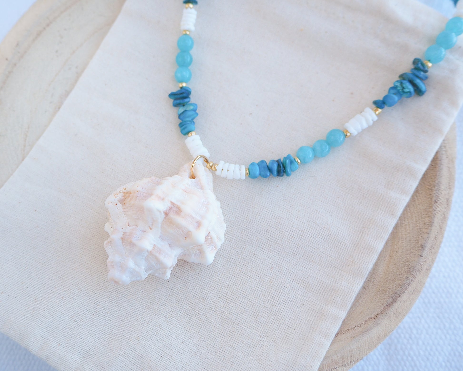 Necklace with blue beads and a shell pendant on a white surface. A necklace with a ivory murex shell paired with aquamarine gemstone beads and white shell beads. The shell is a banded dye murex shell from Portugal and is about 6 cm. The color of the shell is ivory bege.