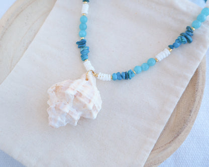 Necklace with blue beads and a shell pendant on a white surface. A necklace with a ivory murex shell paired with aquamarine gemstone beads and white shell beads. The shell is a banded dye murex shell from Portugal and is about 6 cm. The color of the shell is ivory bege.