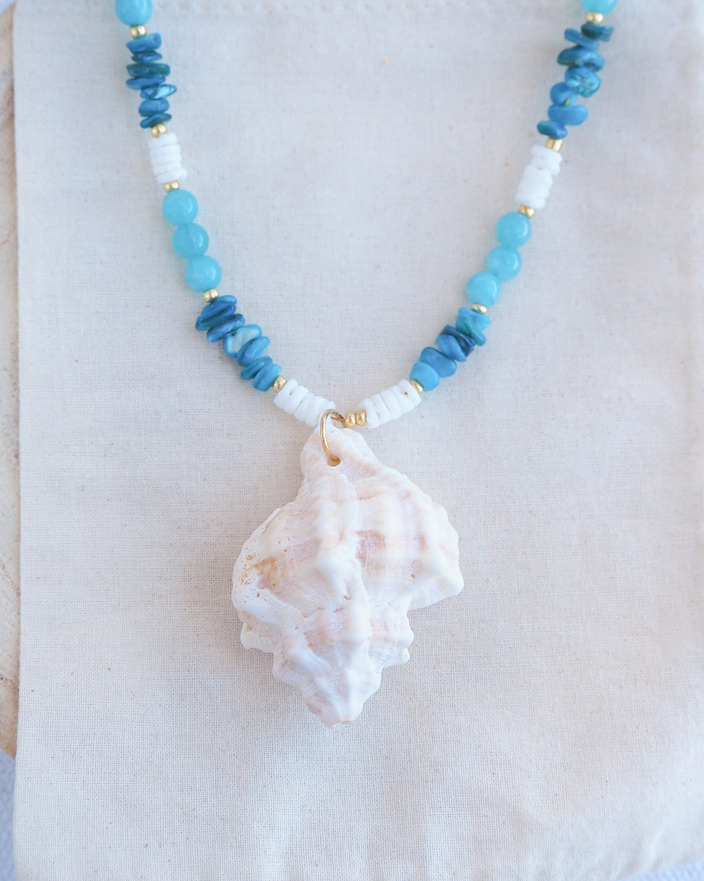 Necklace with blue and white beads and a shell pendant on a white background. Front view of a necklace with a ivory murex shell paired with aquamarine gemstone beads and white shell beads. The shell is a banded dye murex shell from Portugal and is about 6 cm. The color of the shell is ivory bege.