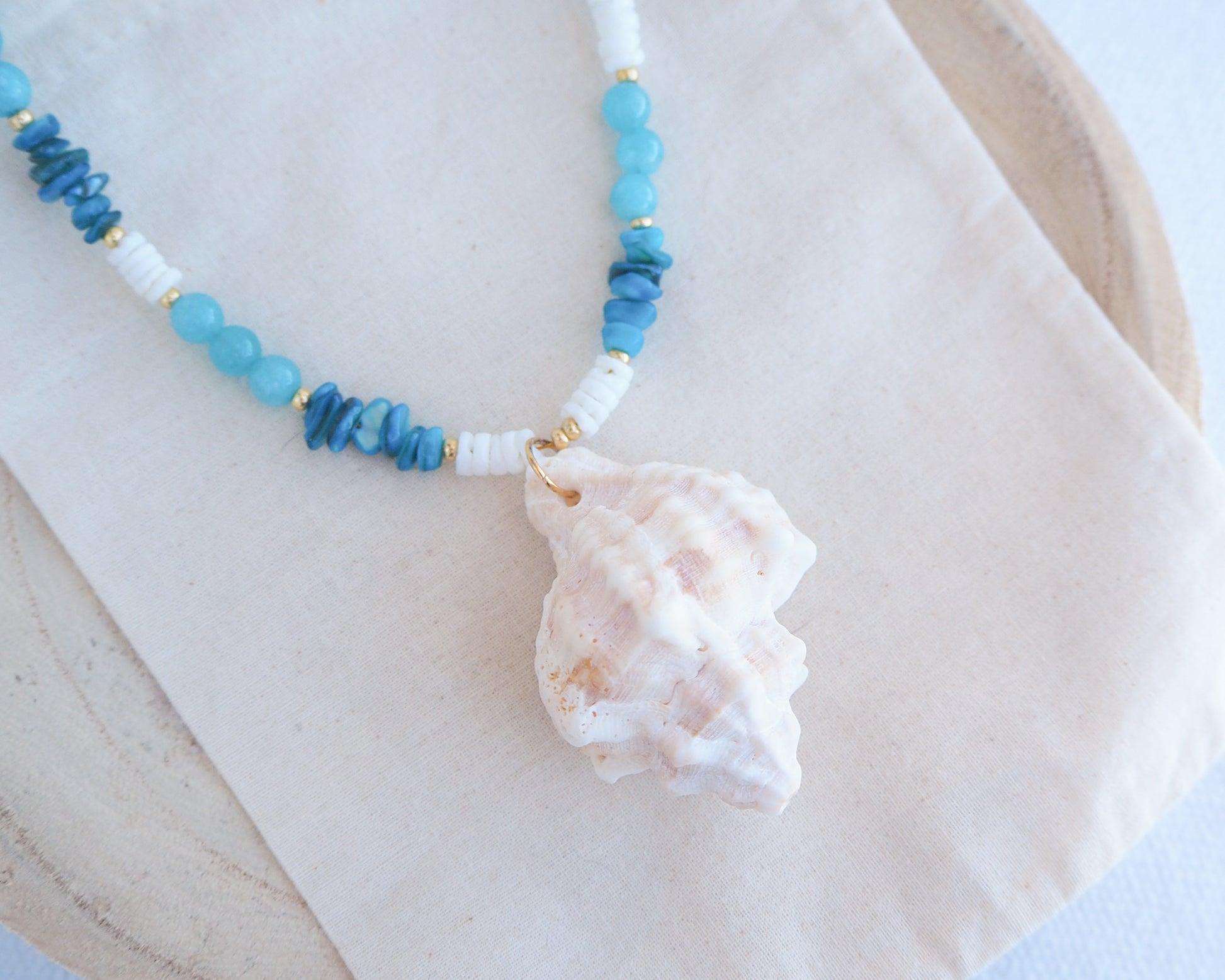 Necklace with blue and white beads and a large shell pendant on a light background. A necklace with a ivory murex shell paired with aquamarine gemstone beads and white shell beads. The shell is a banded dye murex shell from Portugal and is about 6 cm. The color of the shell is ivory bege.