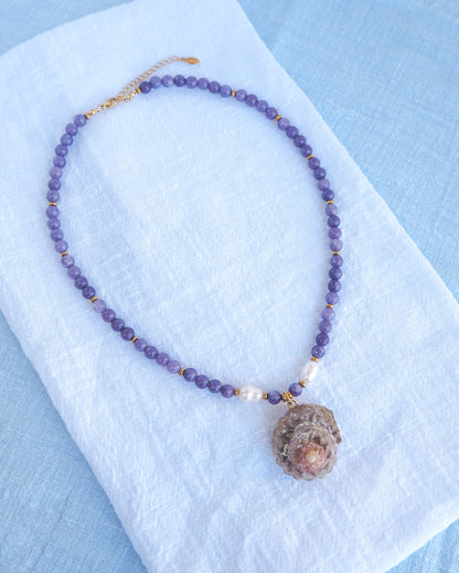 Necklace with purple beads and a large stone pendant on a white cloth with a blue background. 