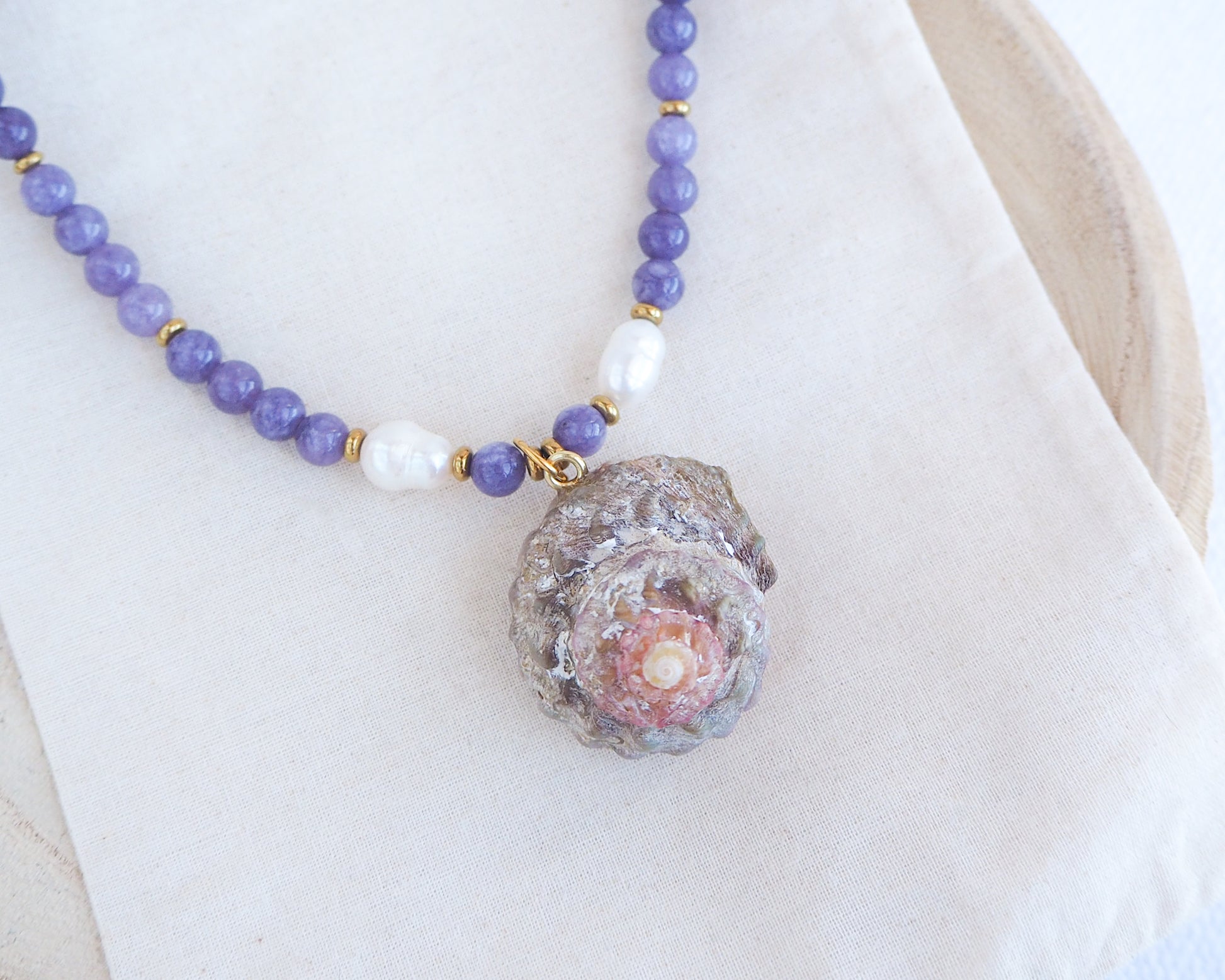 Necklace with purple beads and a textured stone pendant on a white background. Necklace with purple beads and a shell pendant on a white background. A necklace with a big star shell paired with purple amethyst gemstone beads  and freshwater pearls. The shell is a rough star shell from Portugal and is about 4 cm. The color of the shell is kaki grey white.