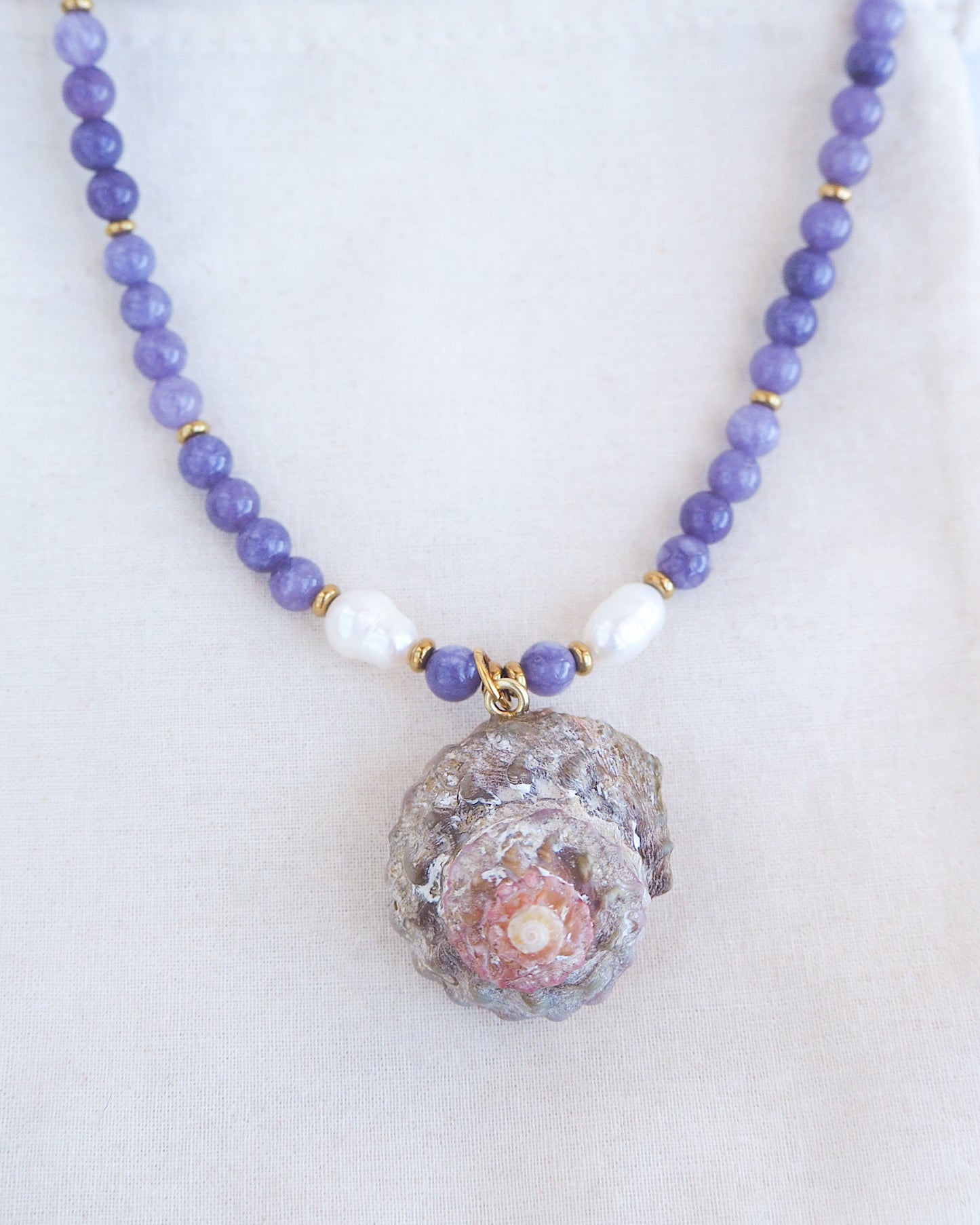 Front view of a necklace with a big star shell paired with purple amethyst gemstone beads  and freshwater pearls. The shell is a rough star shell from Portugal and is about 4 cm. The color of the shell is kaki grey white.