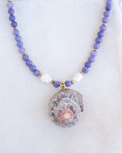 Front view of a necklace with a big star shell paired with purple amethyst gemstone beads  and freshwater pearls. The shell is a rough star shell from Portugal and is about 4 cm. The color of the shell is kaki grey white.