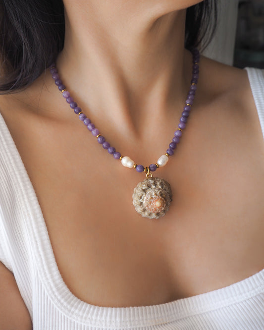 Model wearing a necklace with a big star shell paired with purple amethyst gemstone beads  and freshwater pearls. The shell is a rough star shell from Portugal and is about 4 cm. The color of the shell is kaki grey white.