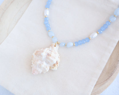 Side view of a necklace with light blue beads and a large shell pendant on a white background. Handmade with a real big banded murex shell handpicked from Portugal, blue lace agate gemstone beads, blue sparkling crystal beads, ivory freshwater pearls and 18k gold stainless steel beads. The necklace is inspired on the ocean is part of the ocean serenity pearl collection. 