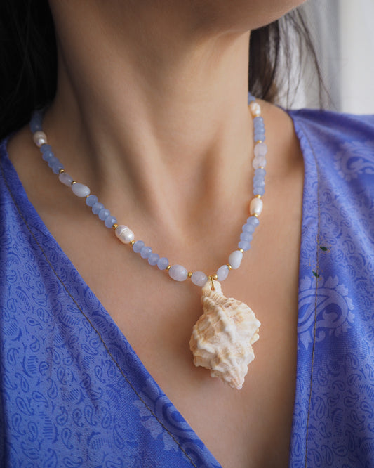 Model wearing a necklace with light blue beads and a large shell pendant on a white background. Handmade with a real big banded murex shell handpicked from Portugal, blue lace agate gemstone beads, blue sparkling crystal beads, ivory freshwater pearls and 18k gold stainless steel beads. The necklace is inspired on the ocean is part of the ocean serenity pearl collection. 