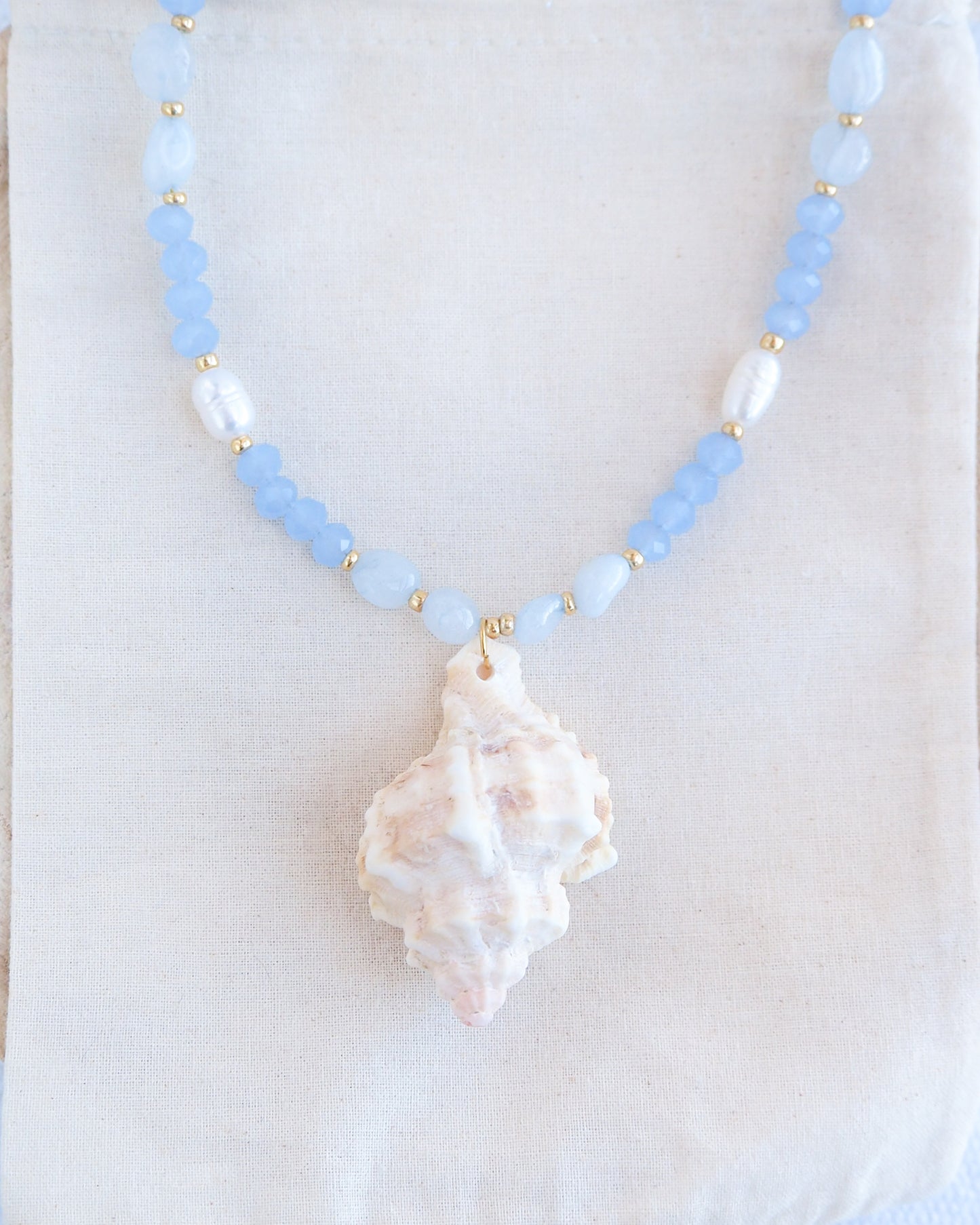 Front view of necklace with light blue beads and a large shell pendant on a white background. Handmade with a real big banded murex shell handpicked from Portugal, blue lace agate gemstone beads, blue sparkling crystal beads, ivory freshwater pearls and 18k gold stainless steel beads. The necklace is inspired on the ocean is part of the ocean serenity pearl collection. 