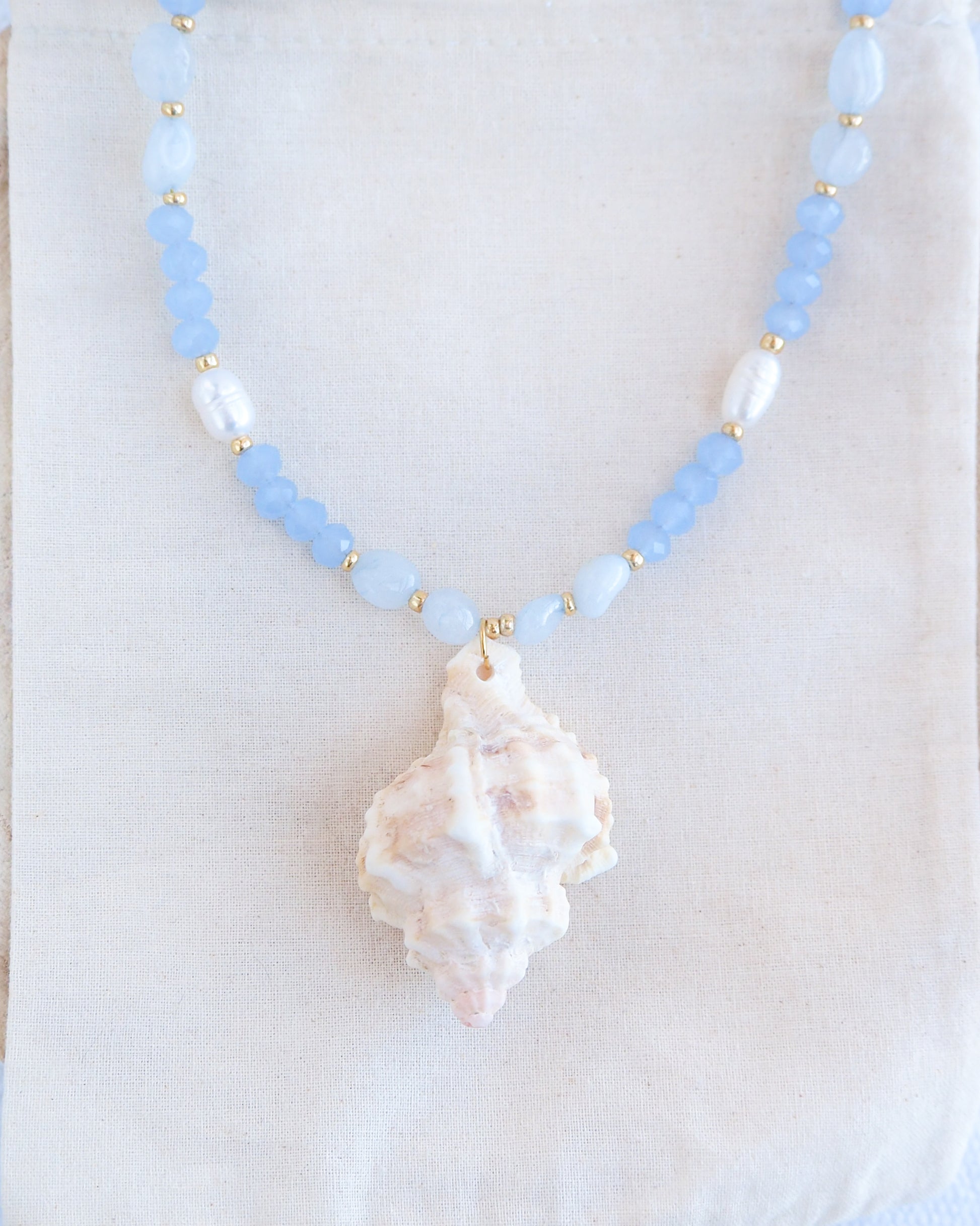Front view of necklace with light blue beads and a large shell pendant on a white background. Handmade with a real big banded murex shell handpicked from Portugal, blue lace agate gemstone beads, blue sparkling crystal beads, ivory freshwater pearls and 18k gold stainless steel beads. The necklace is inspired on the ocean is part of the ocean serenity pearl collection. 
