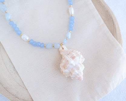 A necklace with light blue beads and a large murex shell pendant on a white background. Handmade with a real big banded murex shell handpicked from Portugal, blue lace agate gemstone beads, blue sparkling crystal beads, ivory freshwater pearls and 18k gold stainless steel beads. The necklace is inspired on the ocean is part of the ocean serenity pearl collection. 