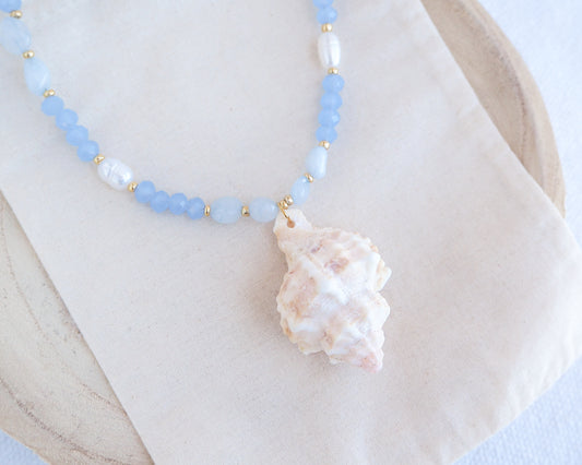 A necklace with light blue beads and a large murex shell pendant on a white background. Handmade with a real big banded murex shell handpicked from Portugal, blue lace agate gemstone beads, blue sparkling crystal beads, ivory freshwater pearls and 18k gold stainless steel beads. The necklace is inspired on the ocean is part of the ocean serenity pearl collection. 