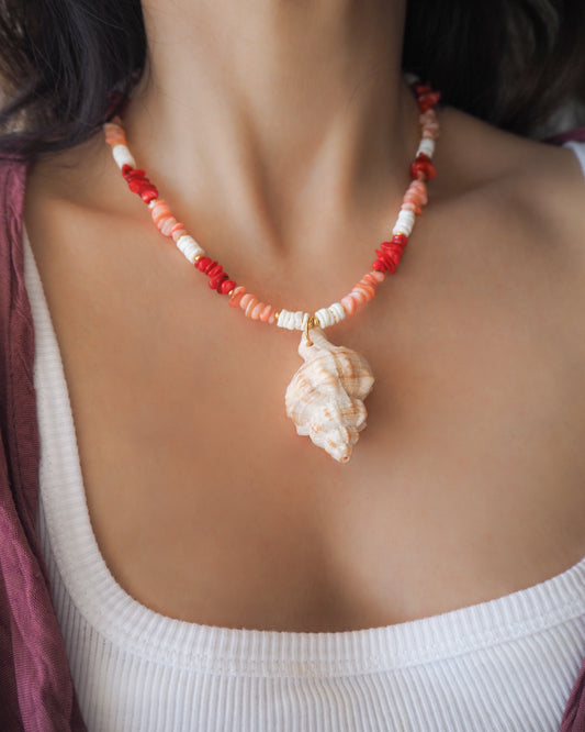 Model wearing a necklace with red, pink, and white beads and a shell pendant on a beige surface. Big murex shell paired with red coral stones and white shell beads. The shell is a banded dye murex shell from Portugal and is about 6 cm. The color of the shell is ivory bege.