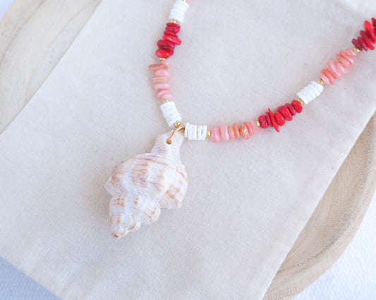 Side view of a necklace with red, pink, and white beads and a shell pendant on a beige surface. Big murex shell paired with red coral stones and white shell beads. The shell is a banded dye murex shell from Portugal and is about 6 cm. The color of the shell is ivory bege.