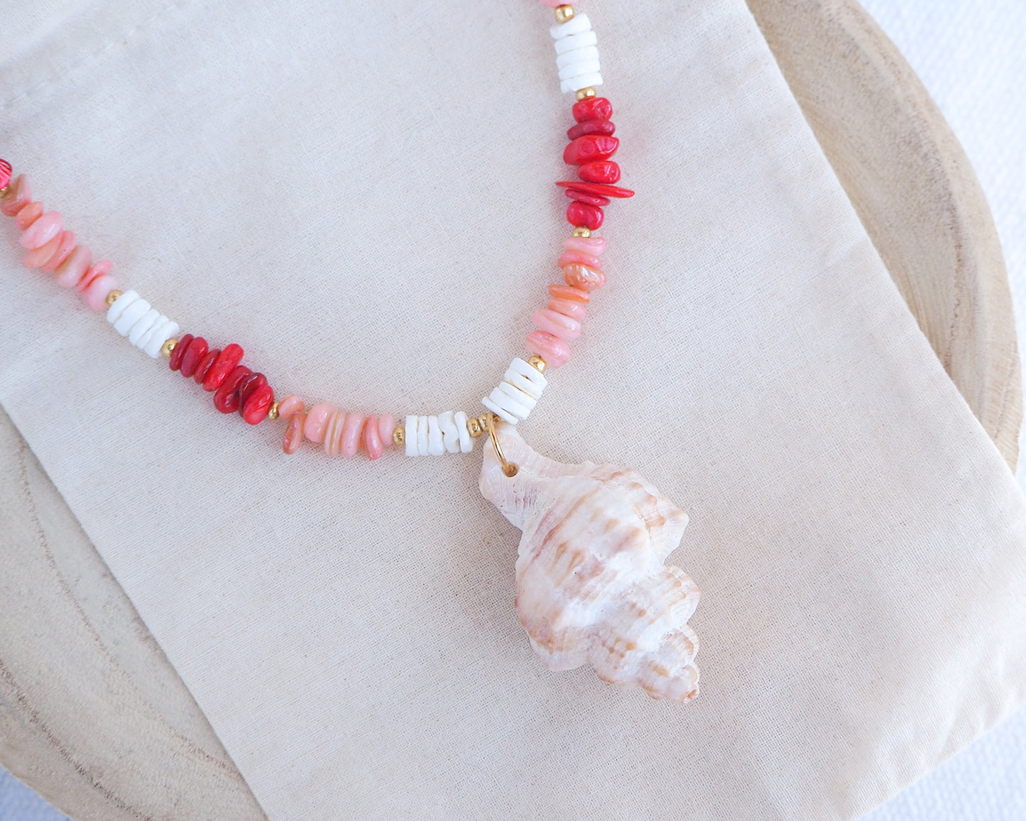 Necklace with red, pink, and white beads and a shell pendant on a beige surface. Big murex shell paired with red coral stones and white shell beads. The shell is a banded dye murex shell from Portugal and is about 6 cm. The color of the shell is ivory bege.