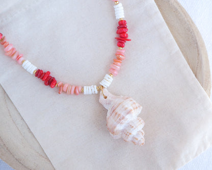 Necklace with red, pink, and white beads and a shell pendant on a beige surface. Big murex shell paired with red coral stones and white shell beads. The shell is a banded dye murex shell from Portugal and is about 6 cm. The color of the shell is ivory bege.