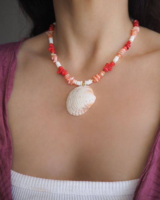 Necklace with shell pendant and red and white beads on a person wearing a purple top. Model wearing a necklace with red, pink, and white beads and a shell pendant on a beige surface. Big venus shell paired with red coral stones and white shell beads. The shell is a Warty Venus Clam shell from Portugal and is about 6 cm. The color of the shell is ivory bege.