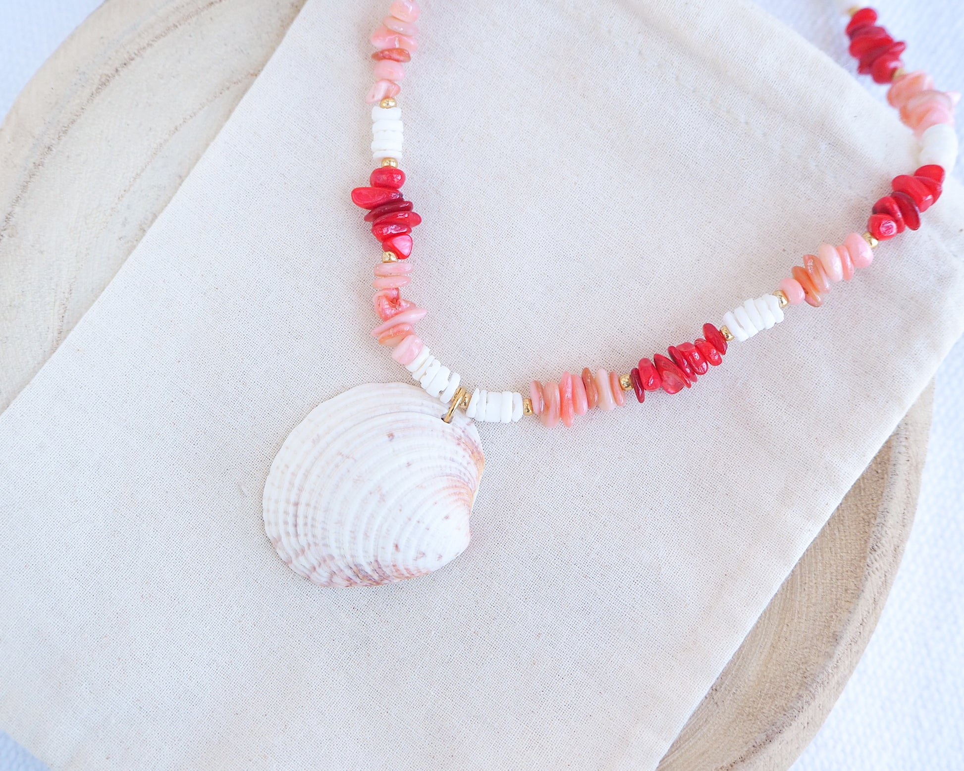 Necklace with shell pendant and coral-colored beads on a white fabric background. a necklace with red, pink, and white beads and a shell pendant on a beige surface. Big venus shell paired with red coral stones and white shell beads. The shell is a Warty Venus Clam shell from Portugal and is about 6 cm. The color of the shell is ivory bege.