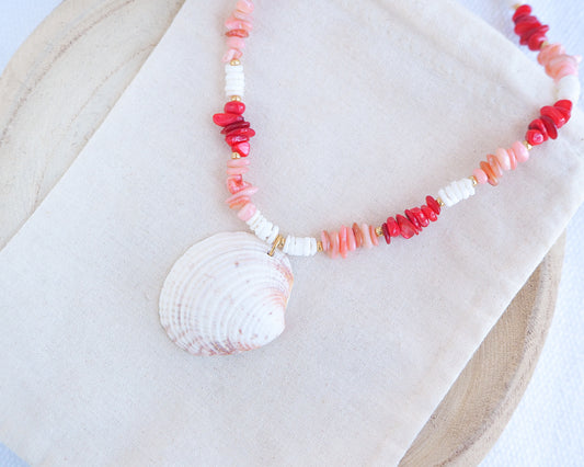 Necklace with shell pendant and coral-colored beads on a white fabric background. a necklace with red, pink, and white beads and a shell pendant on a beige surface. Big venus shell paired with red coral stones and white shell beads. The shell is a Warty Venus Clam shell from Portugal and is about 6 cm. The color of the shell is ivory bege.