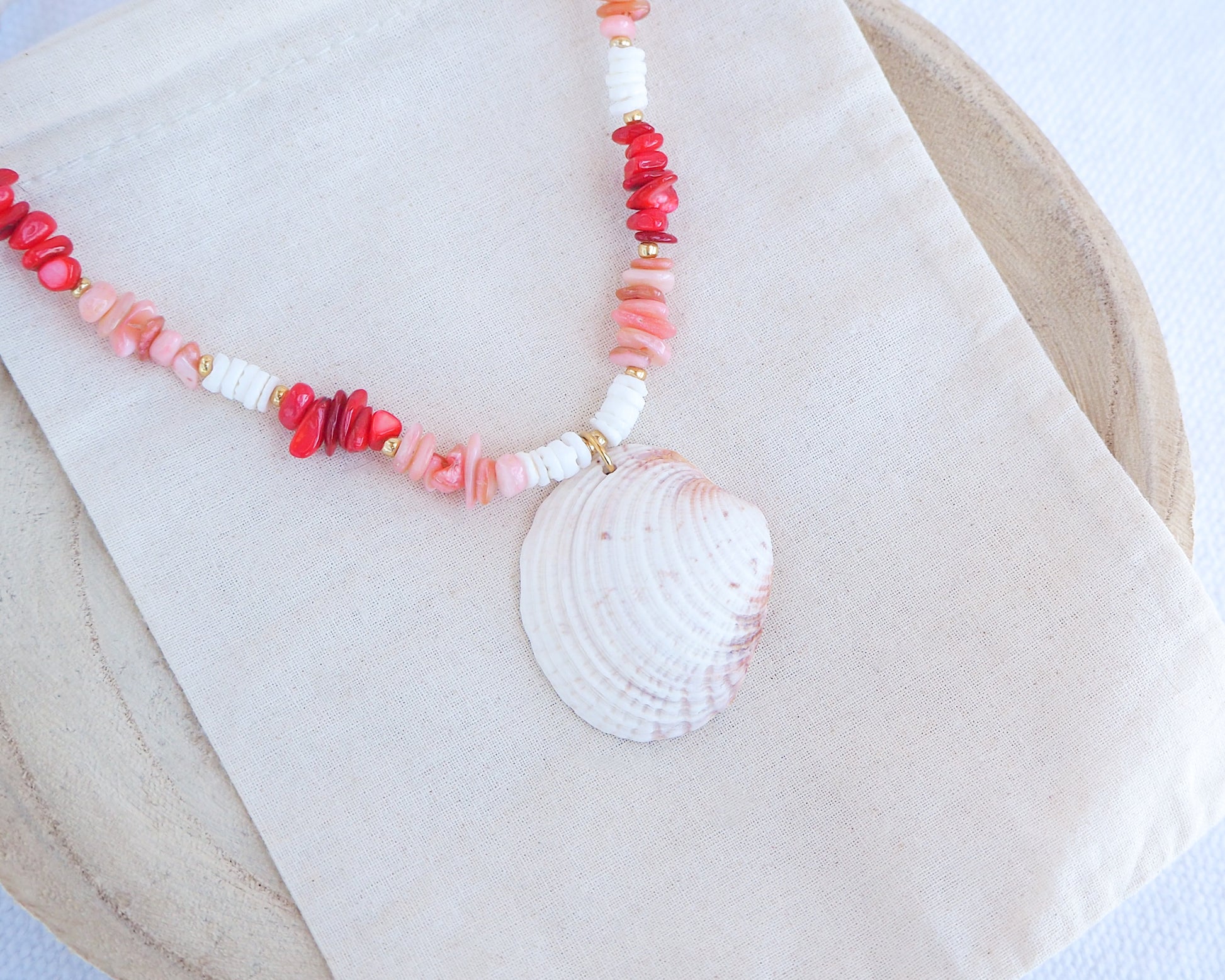 Side view of Necklace with shell pendant and coral-colored beads on a white fabric background. a necklace with red, pink, and white beads and a shell pendant on a beige surface. Big venus shell paired with red coral stones and white shell beads. The shell is a Warty Venus Clam shell from Portugal and is about 6 cm. The color of the shell is ivory bege.