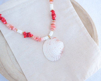 Side view of Necklace with shell pendant and coral-colored beads on a white fabric background. a necklace with red, pink, and white beads and a shell pendant on a beige surface. Big venus shell paired with red coral stones and white shell beads. The shell is a Warty Venus Clam shell from Portugal and is about 6 cm. The color of the shell is ivory bege.