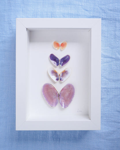 Framed butterfly decorations on a blue fabric background. Artwork with butterfly-shaped seashells in various colors on a light gray background. Made with handpicked shells from Portugal. Coquina shells, thin tellin shells, and rayed trough shells. Set in a white frame box.