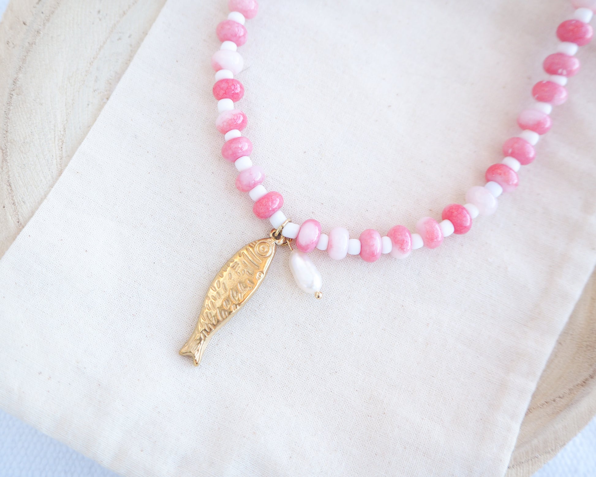 Side view of a necklace with pink and white beads and a gold sardine fish pendant and freshwater pearl bead on a person wearing a white top. The necklace is made with seed beads and freshwater pearls and a big gold sardine fish The sardine fish pendant is made of 18k gold stainless steel. 