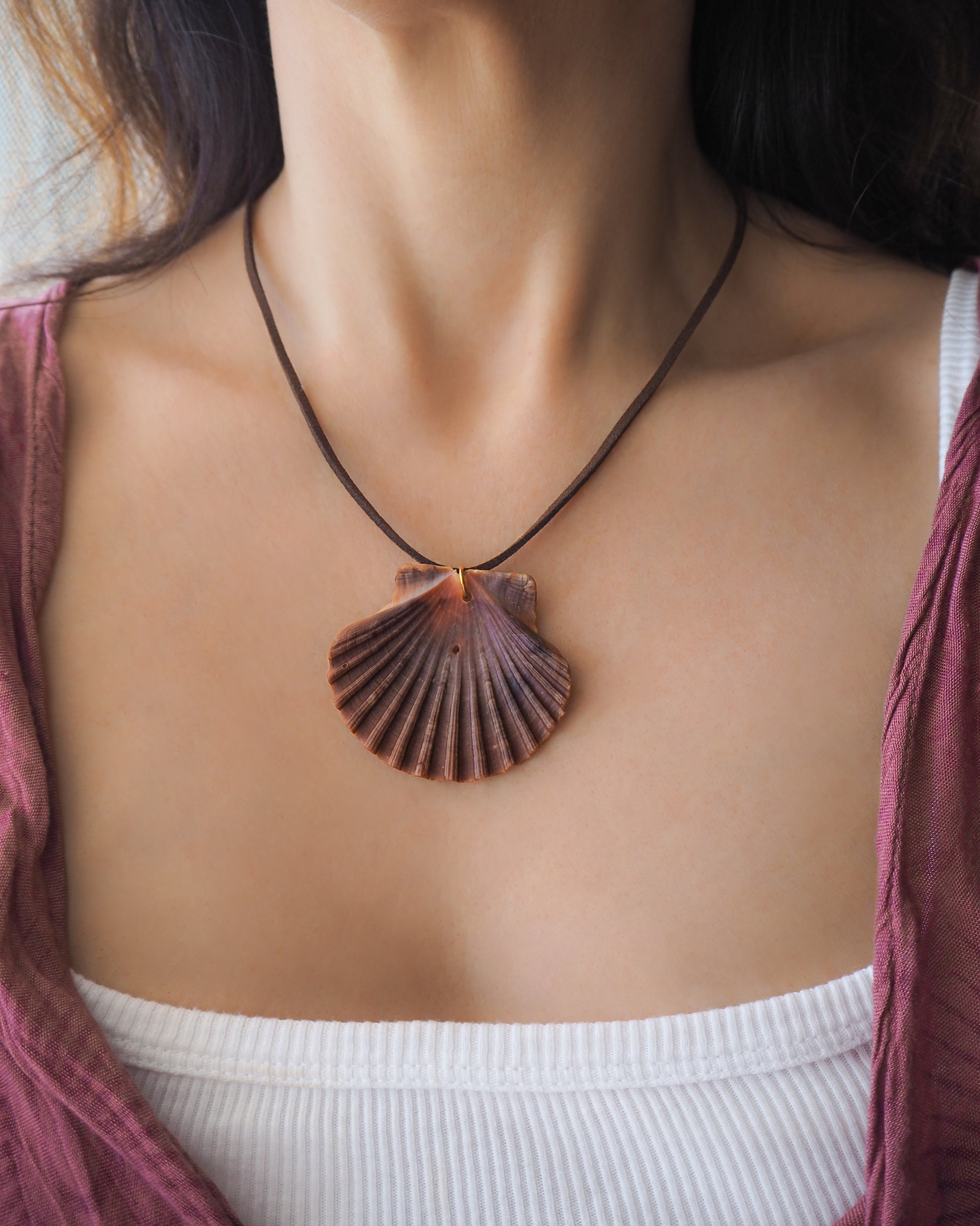 Model wearing a necklace with a big scallop shell on a brown suede cord. The shell is a great scallop shell from Portugal and is about 6 cm. The color of the shell is bordeaux brown and it has vertical ridges. 