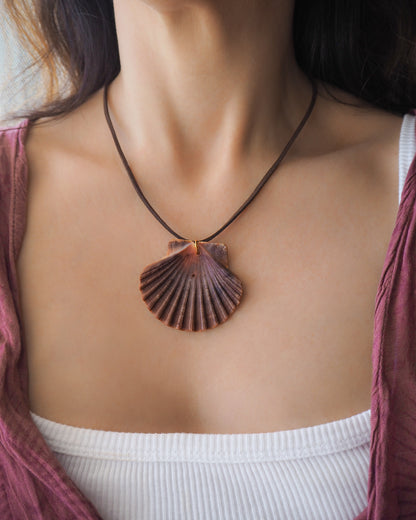 Model wearing a necklace with a big scallop shell on a brown suede cord. The shell is a great scallop shell from Portugal and is about 6 cm. The color of the shell is bordeaux brown and it has vertical ridges. 